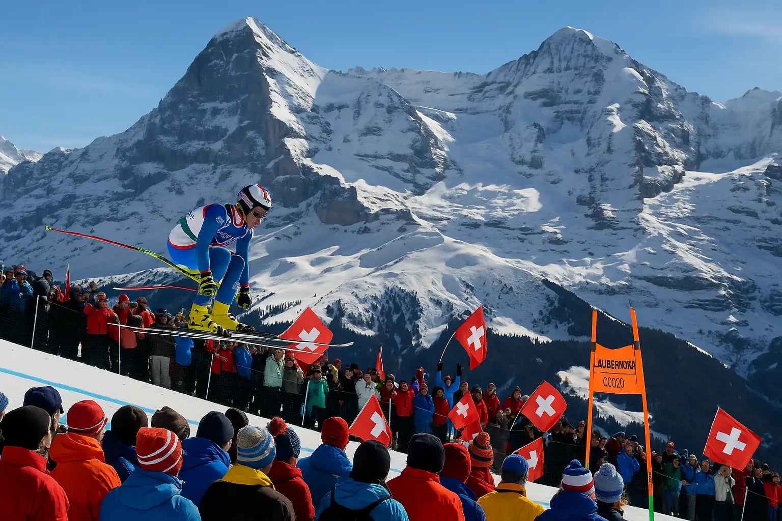 Skirennfahrer springt beim Lauberhornrennen in der Schweiz vor jubelndem Publikum mit Schweizer Fahnen, im Hintergrund die verschneiten Gipfel von Eiger, Mönch und Jungfrau – Lauberhornrennen Schweiz Skisport Alpen