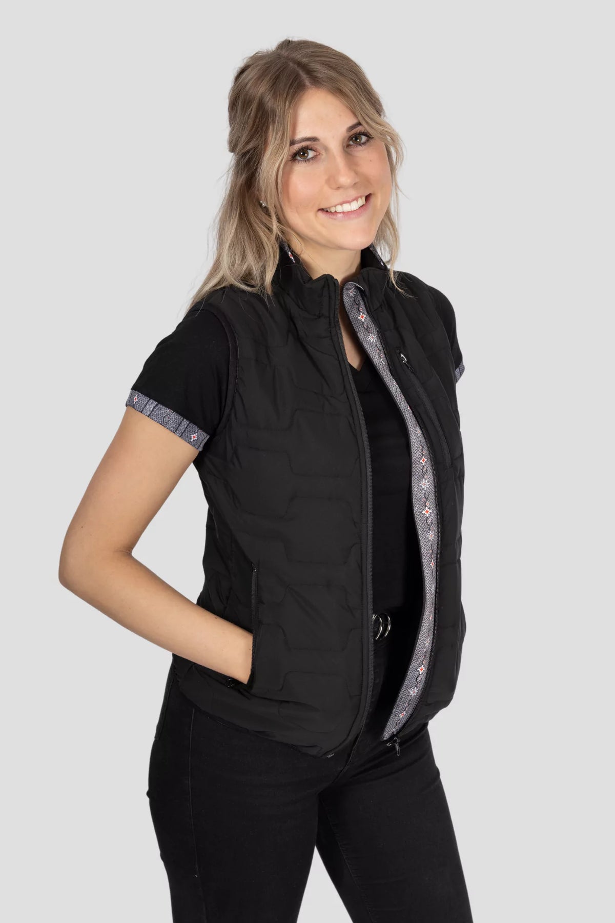 Gilet Edelweiss Damen in Schwarz mit grauem Edelweiss-Muster, hochwertig verarbeitet, mit dezenten Edelweiss-Streifen am Reissverschluss und Nacken, ideal für Freizeit, Arbeit und elegante Anlässe